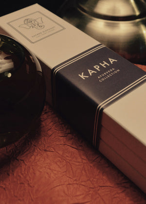 A box of KAPHA incense sticks with a stick partially burned, placed on a red surface, with a small amount of smoke rising.