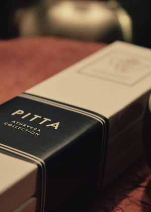 A box labeled 'PITTA' from the Ayurveda Collection, with incense sticks visible to the side, placed on a red surface.